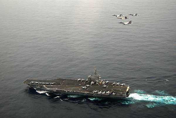 An F/A-18E Super Hornet, an F-22A Raptor, an F-15C Eagle, and an F/A-18C Hornet fly in formation above USS Ronald Reagan (CVN 76).
